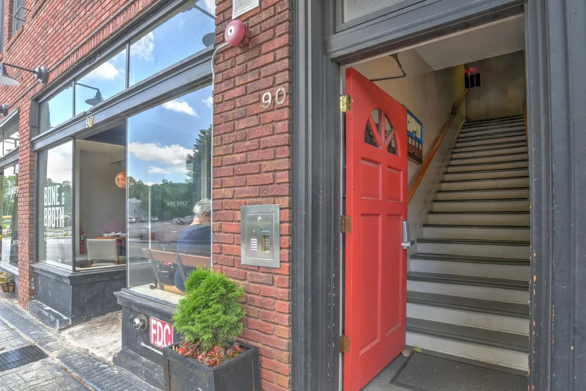 This image captures the welcoming exterior of a building with a vibrant red door set in a historic brick facade, complemented by large windows. The scene suggests an inviting entry point, likely a café or retail spot, with people potentially stepping in, enjoying the lively street atmosphere or pausing to people-watch.