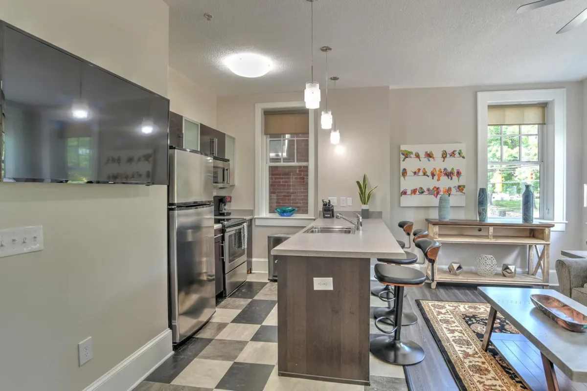 This image centers on the renovated kitchen, showcasing a stylish kitchen island, sleek stainless steel appliances, modern artwork on the wall, bar stools for casual dining, and a window allowing natural light to flood the space. The scene suggests a harmonious setting for both preparing meals and entertaining guests.