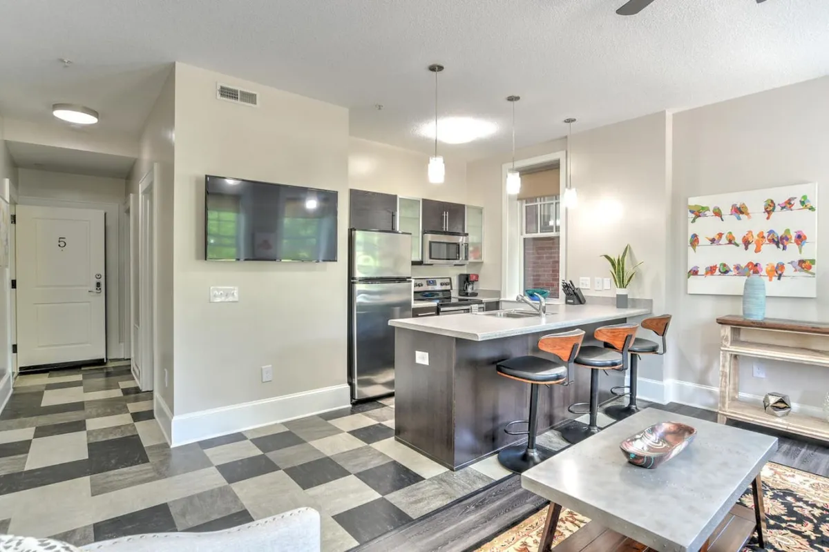 The image showcases the open-concept kitchen and living area of a stylishly renovated condo. A spacious kitchen island with modern bar stools invites conversation, while vibrant artwork and a large television set a cozy, contemporary tone. The space is bright and welcoming, ideal for both casual hangouts and festive gatherings.