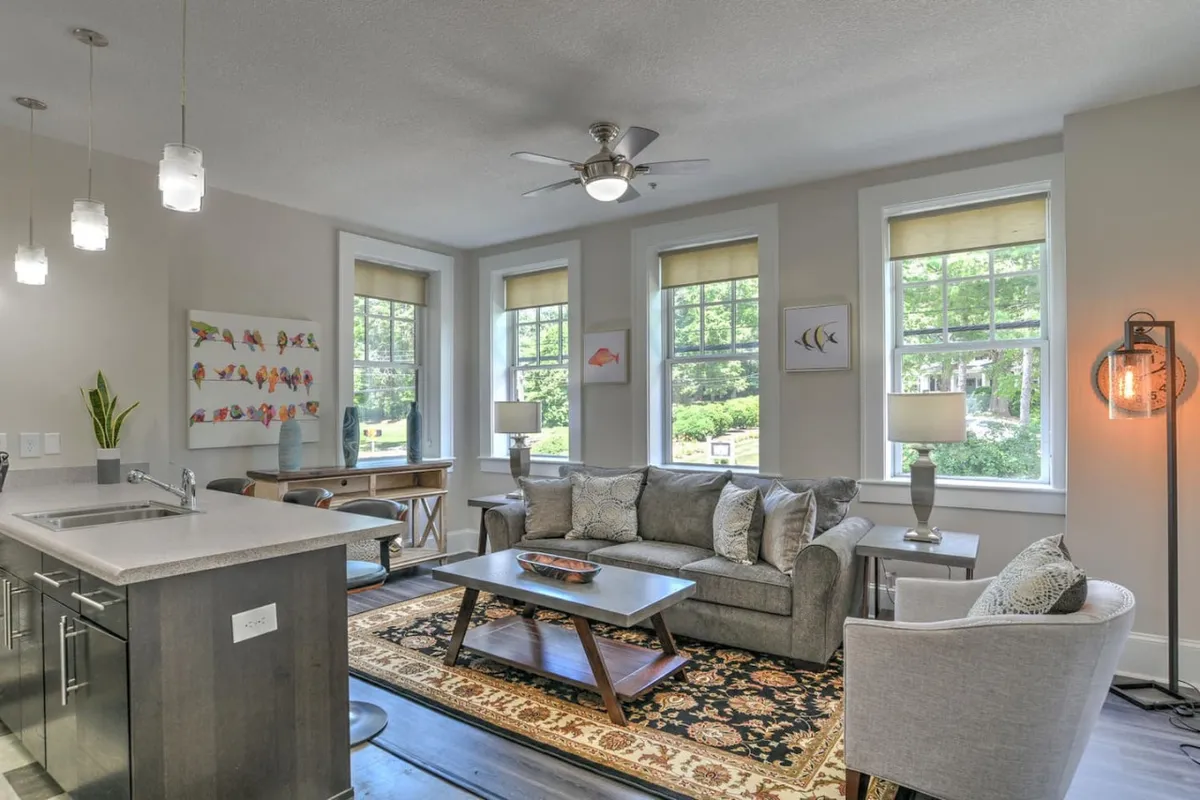 A sunlit living room featuring a comfortable sofa, coffee table, decorative art on the walls, and large windows that flood the space with natural light. The room has a welcoming, open feel, with original architectural details visible and a boutique, stylish ambiance.
