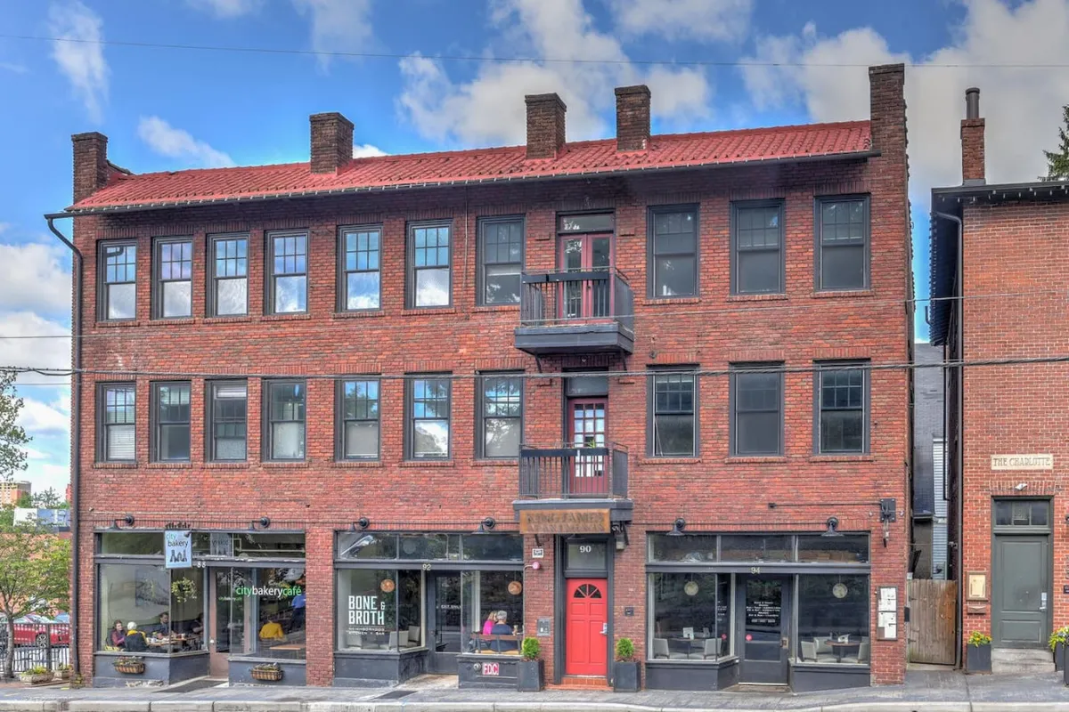 A warmly lit brick building featuring a distinctive red door, upper balcony, and bustling storefronts below. The scene suggests an energetic neighborhood where locals and visitors enjoy outdoor seating, explore unique shops, and connect with the community.