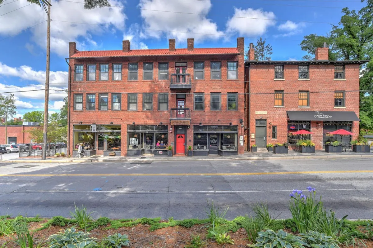 This exterior image captures a lively urban street scene featuring a stately brick facade, an inviting red door, a balcony, and an outdoor seating area. The atmosphere is vibrant with hints of dining, shopping, and socializing, reflecting the energy of North Asheville's community spaces.